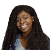 A person with long curly hair is smiling while wearing a gray shirt and a denim jacket.