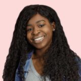 Smiling person with long curly hair wearing a denim jacket over a gray shirt against a pink background.