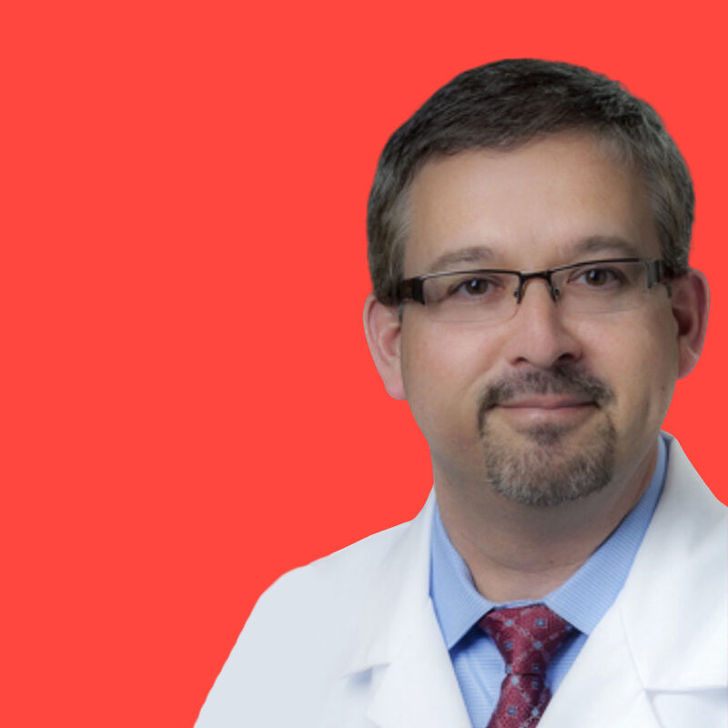 A person wearing glasses, a white coat, a blue shirt, and a red tie stands against a solid red background.