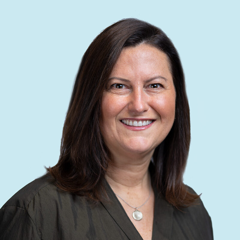 A person with shoulder-length brown hair and a dark green top smiles at the camera against a light blue background.