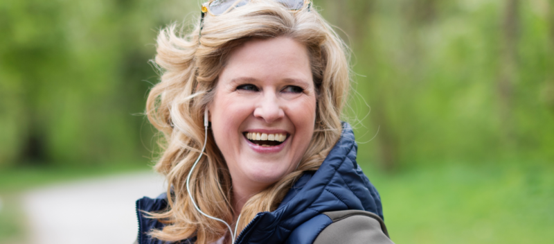 Person with wavy blonde hair, wearing a blue vest, smiling outdoors with greenery in the background.