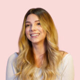 Woman with long wavy blonde hair smiling, wearing a light-colored off-the-shoulder top, against a pink background.