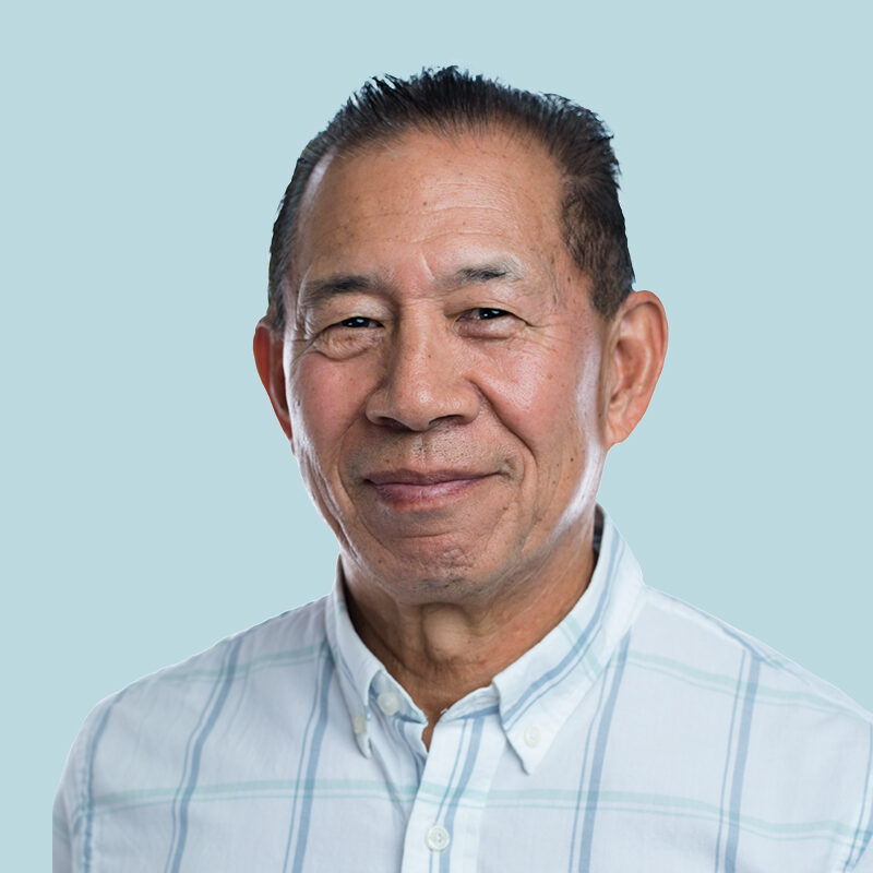 A person with short dark hair is smiling slightly, wearing a light plaid shirt, set against a pale blue background.