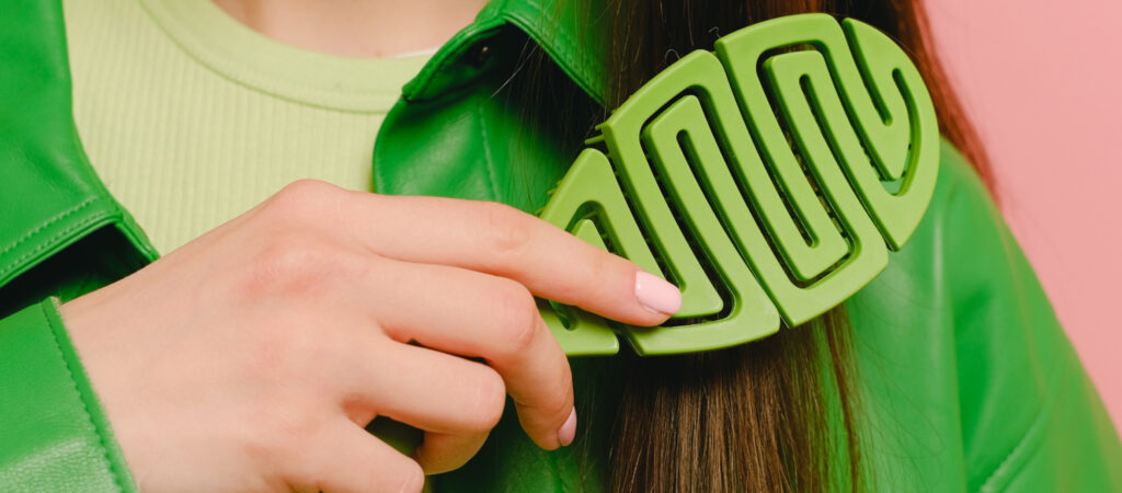 A person with long brown hair holds a large green hair clip against a green jacket.