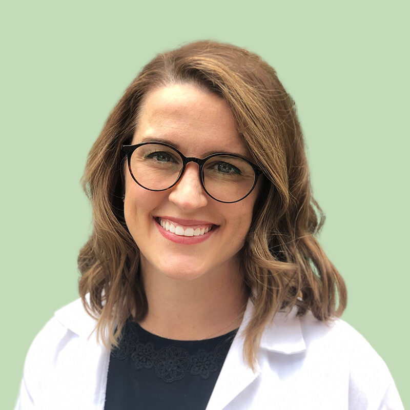 A person with brown hair and glasses smiles, wearing a white lab coat against a light green background.