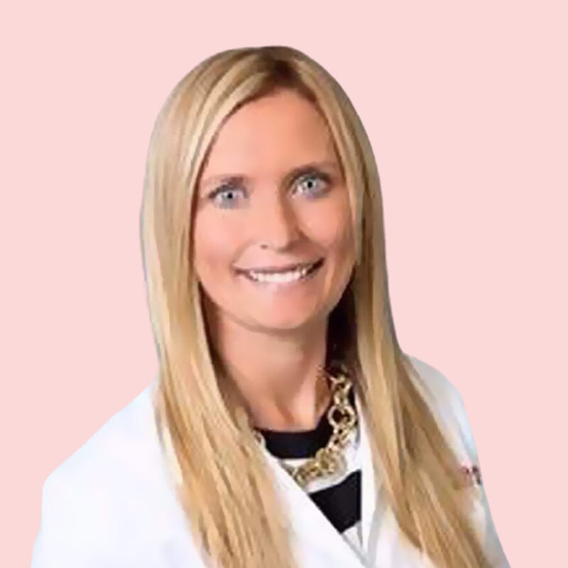 Person with long blonde hair in a white coat smiles against a plain pink background.