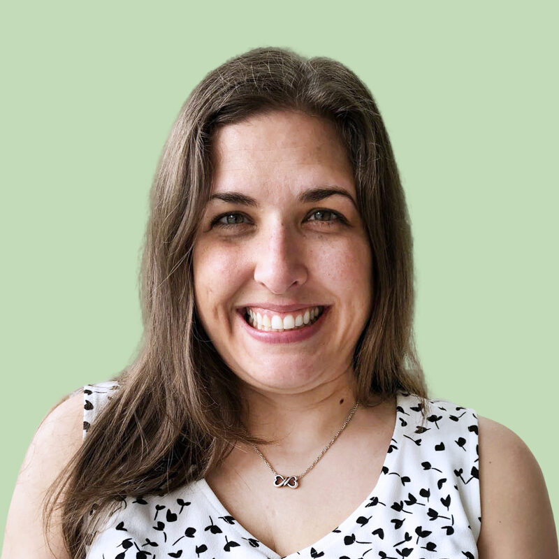 Smiling person with long brown hair wearing a white and black patterned shirt stands in front of a light green background.