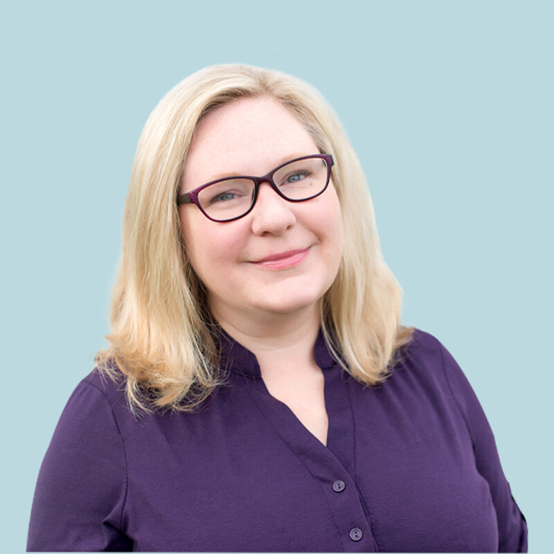 A person with blonde hair and glasses is wearing a purple shirt against a light blue background.